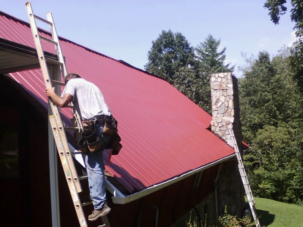 Roofer on a ladder beside a red metal roof and stone chimney — Soffit & Fascia Repair by Roofing Brothers in Samaria